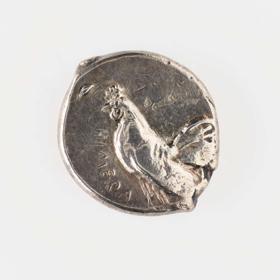 Silver coin with irregular borders. Embossed on one side is the side profile of a rooster surrounded by Greek lettering.