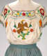 White blouse with a central embroidered eagle atop a green cactus framed by the Mexican flag and colorful detailed floral patterns tucked into a blue cinched skirt waistband. 
