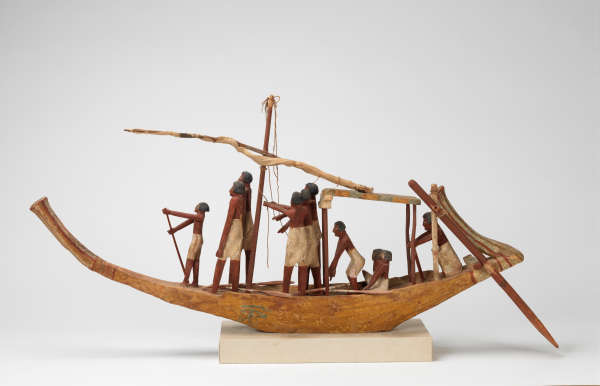 A carved wooden boat model on a rectangular off-white base with a tied up sail manned by sailors with brown skin and white trousers holding oars and ropes.