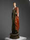 A tall, weathered wooden statue of a robed figure with a serene expression, draped in green and red garments, standing upon an octagonal base.