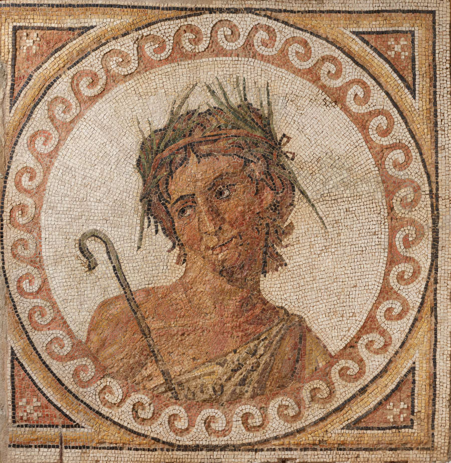 A clay, cream, and green mosaic of a man in a toga wearing a wreath, holding a shepherd’s staff. His portrait is framed by a circle of clay-red swirling patterns.