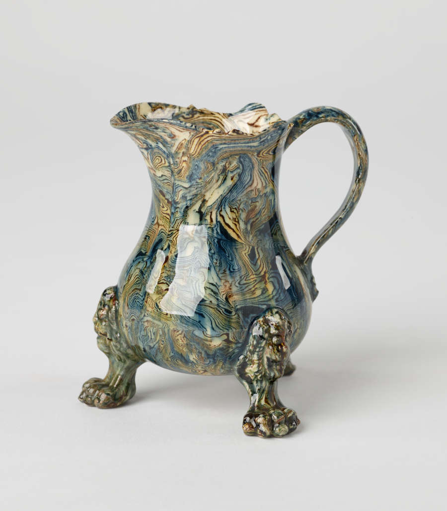 A small vessel with a cream, blue, and green mottled design. Three sculptural feet are shaped like lion’s paws with lion heads attached to body of vessel.