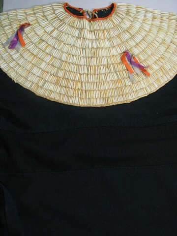 Closeup of the off-white collar of a black dress, which features concentric rows of thread with two orange and purple tassels. The neckline is thin and orange, with a gray-green tie. 