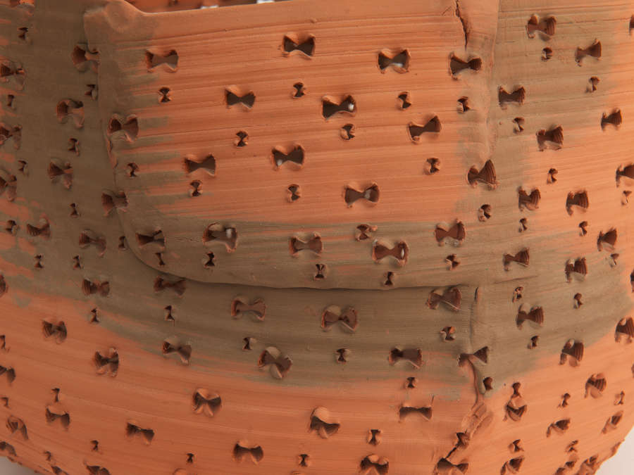 Close-up of a terracotta vessel with a brushed gray wash, its matte surface punctured with repeating bow-shaped holes arranged in slightly uneven rows that cover the whole vessel. 