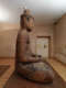 A side view of a seated wooden Buddha statue in a warmly lit room, with visible signs of age and repair. 