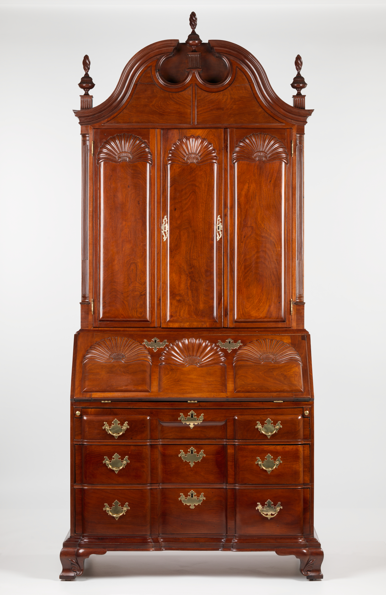 A tall mahogany cabinet and desk structure with a polished finish, slant-front design, intricate shell-shaped carvings, brass keyholes on all drawers, and a grand arched top decorated with ornamental details.