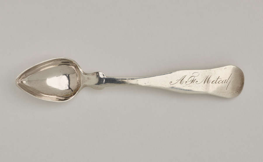 A silver salt spoon the bowl comes to a pointed tip, the neck is thin, and the handle is thicker and rounded at the end.
