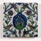 White square tile painted with a dense symmetrical floral pattern in blues, blacks and greens. A large maned flower occupies the center, with smaller flowers of varying shapes surrounding it.