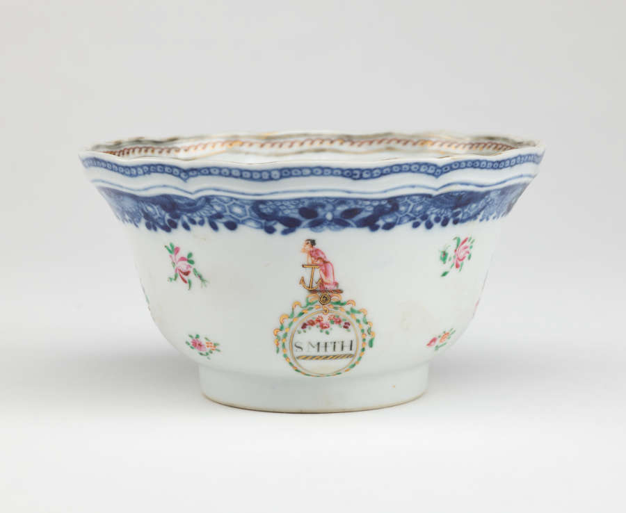 A bowl with glided, blue, green, and pink decorations. The body is decorated with flowers and a figure resting on an anchor. Word below the figure says “SMITH.”
