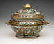 An oval lidded vessel with a large gold finial. The body of the vessel is decorated with multicolored butterflies on a white ground.
