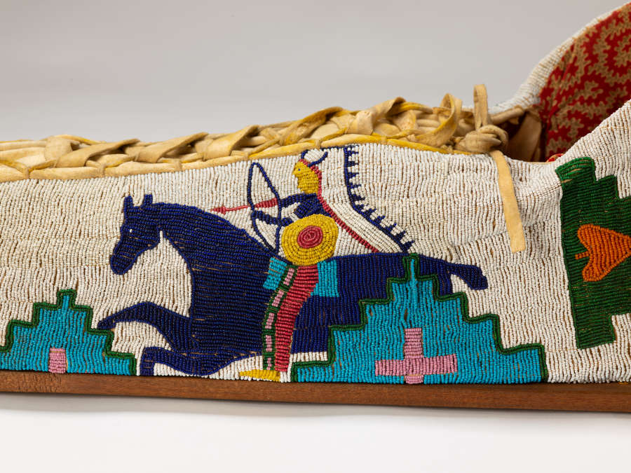 Detail of beaded object. White background beaded with blue horse and colorful rider in headdress, shooting an arrow. Geometric decoration at bottom. Leather lacing at top.