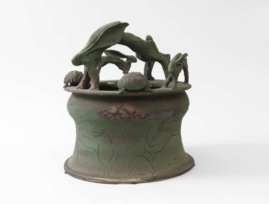 An earthy green sculptural vessel with rabbits and turtles leaping and circling around the rim. The body is lightly etched with flowing line drawings of rabbits in motion.