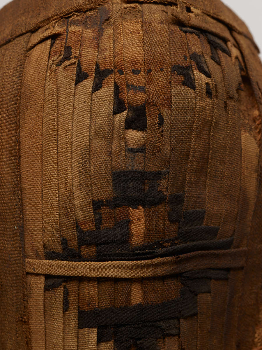 Detail of the long brown almond-shaped woven object, showing the intricacies of its weaving, consisting of various shades of brown cloth coming together and dyed to form geometric diamond patterns.