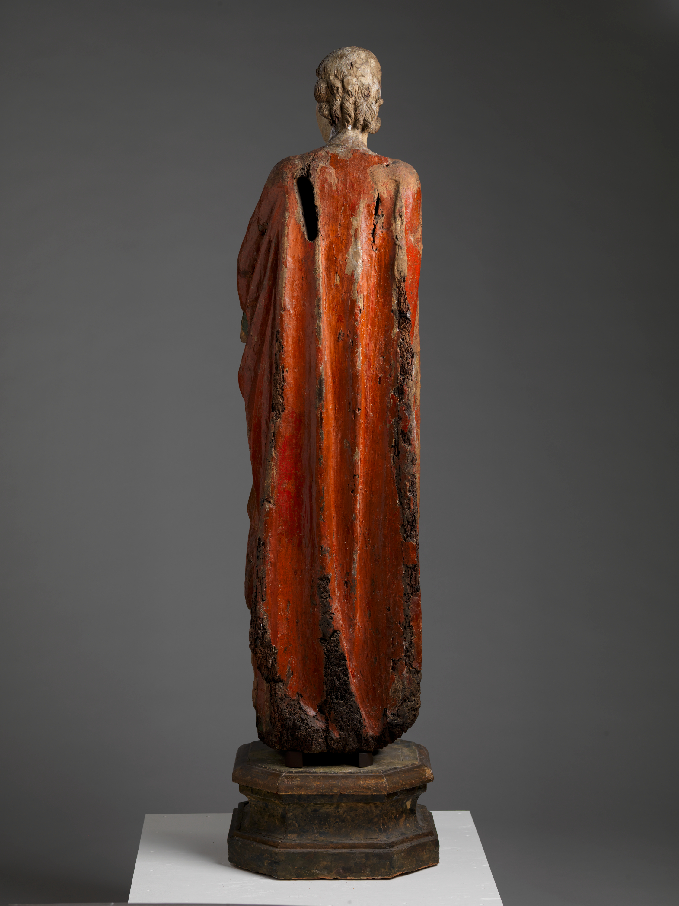 A wooden statue viewed from the rear, draped in a long, faded red robe with visible signs of age with cracks and missing paint.