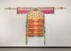A garment displayed on a wooden pole, with bright woven geometric patterns, dangling gourds, and repeated pink text reading "STAND YOUR GROUND" woven into the textile.