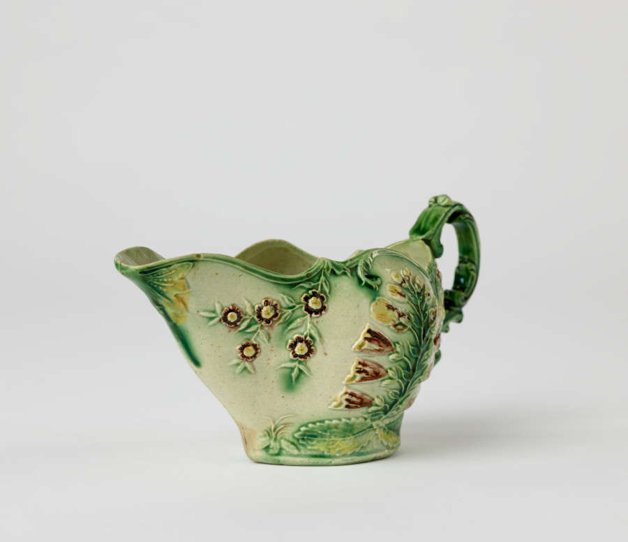  A short boat shaped pitcher green, cream, and brown in color with a sculptural handle and floral decorations.
