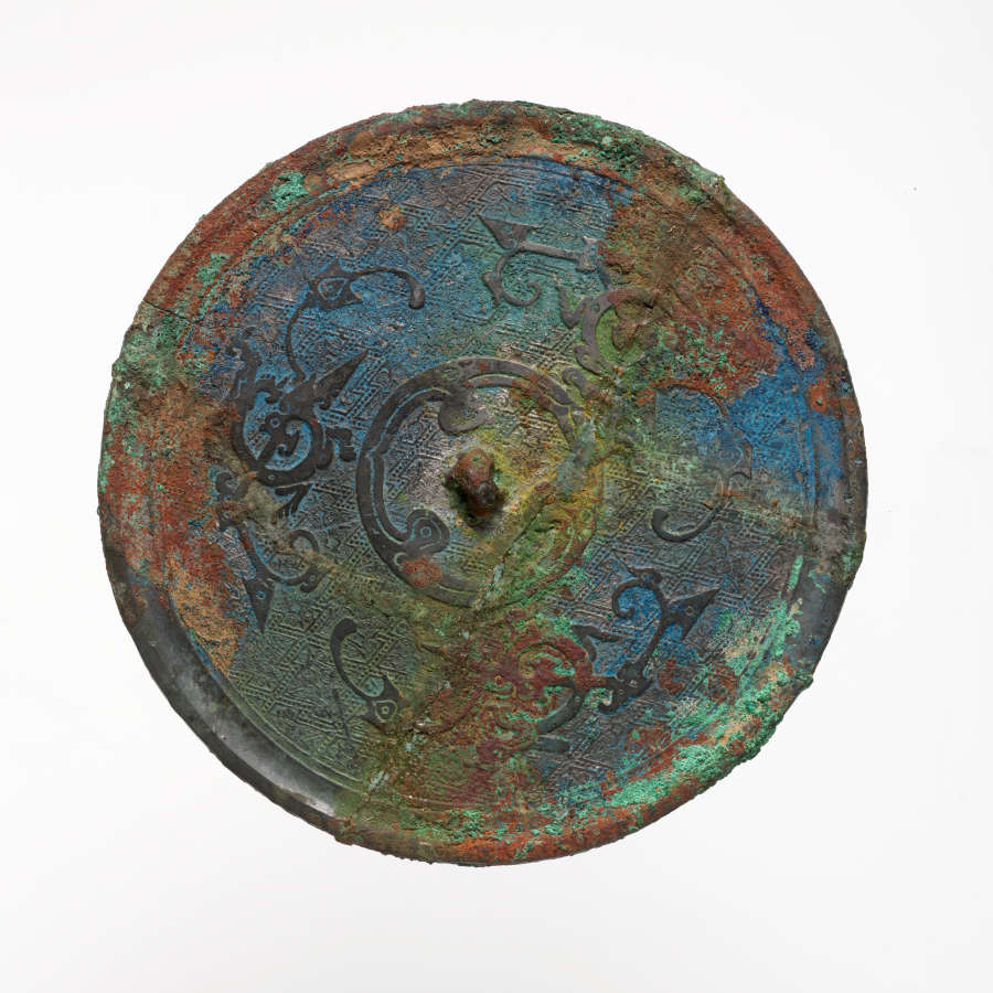 Back of an ornate circular mirror with raised swirls arranged concentrically. Its surface is so worn that it’s no longer reflective, instead consisting of blues, greens, and browns.