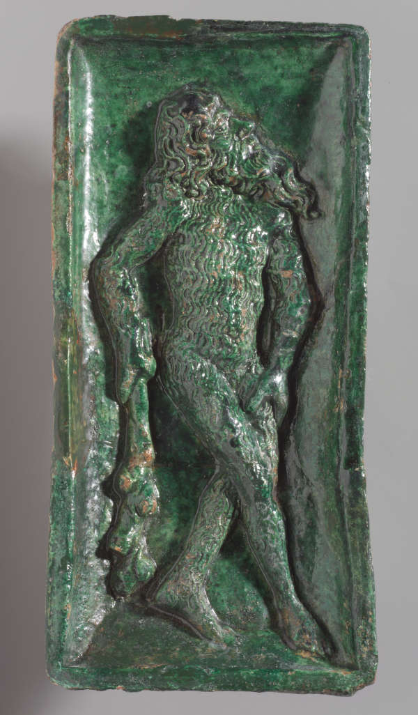 A green sculpted tile of a figure with long hair and beard, holding a stick-like object.