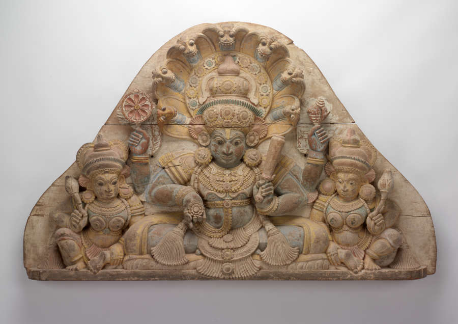 Rounded, cream-colored, triangular carving of three seated figures wearing ornate clothing and headdresses. The central figure is the largest and has four arms, each holding a different object.