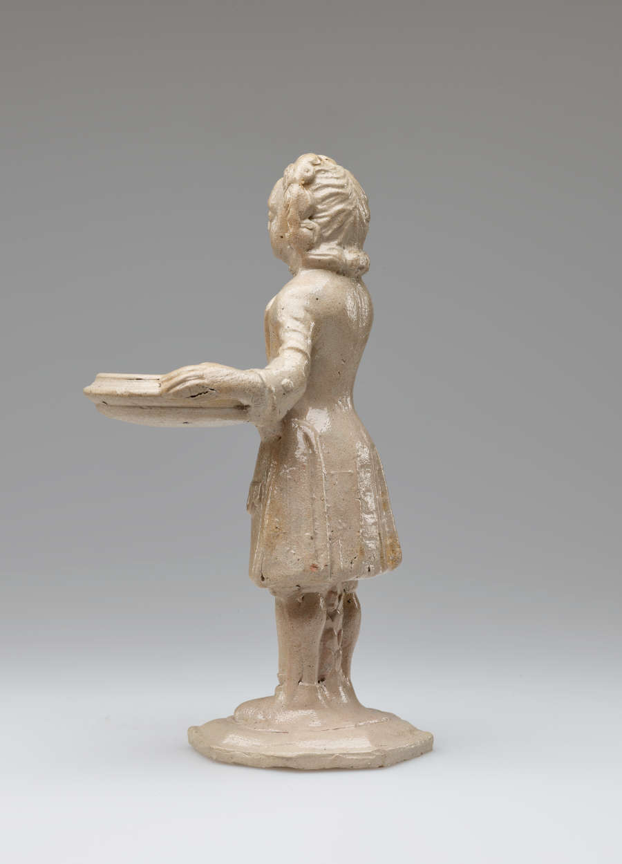 A cream colored sculptural figure holding a vessel, dressed in historical clothing with tightly curled hair.
