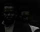 A dark, low contrast black-and-white photograph capturing two nearly imperceptible dark-skinned men gazing at the viewer with inscrutable expressions. One is wearing glasses and partially obscured behind the other. 