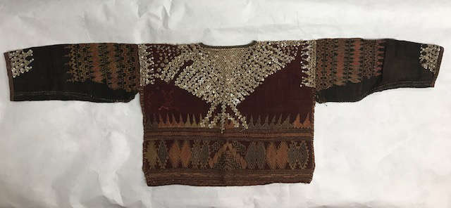 Handcrafted garment in tones of black, brown and deep red on a white background. Geometric designs cover the shoulders and lower chest, while a large light V-shape borders the neckline.