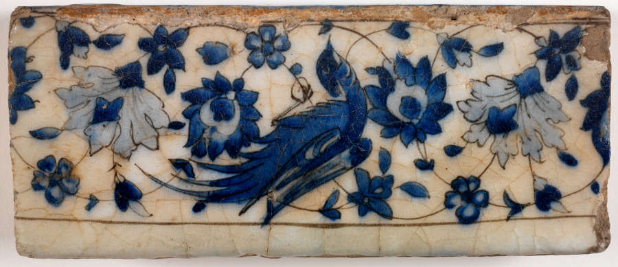 A horizontally long rectangular tile fragment. The top is worn, brown and off-white with indigo designs of a bird against a symmetrically arranged floral pattern. 