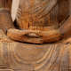 A close-up of a wooden Buddha statue’s hands resting in its lap, with visible wear and erosion.