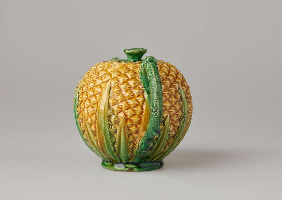 A teapot in the shape of a pineapple. Handle, foot, lid, and spout are green.