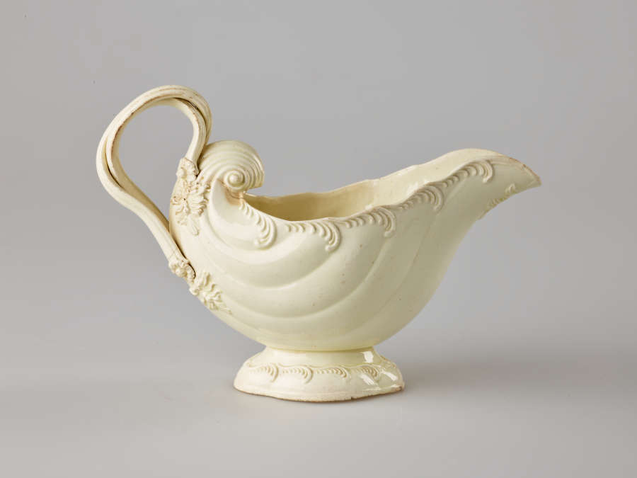  A cream colored vessel with rounded foot and handle. The body of the vessel is shell shaped and has swirled decorations.