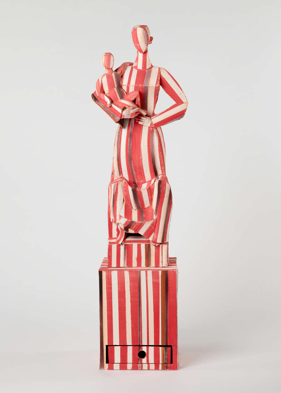 A sculpture of a woman holding a child on top of a rectangular base, covered in red, white, and brown stripes.