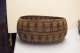Side-view of an ochre-brown oval woven basket in storage, with convex sides featuring two patterned stripes of dark-brown figures holding hands separated by horizontally running stripes.