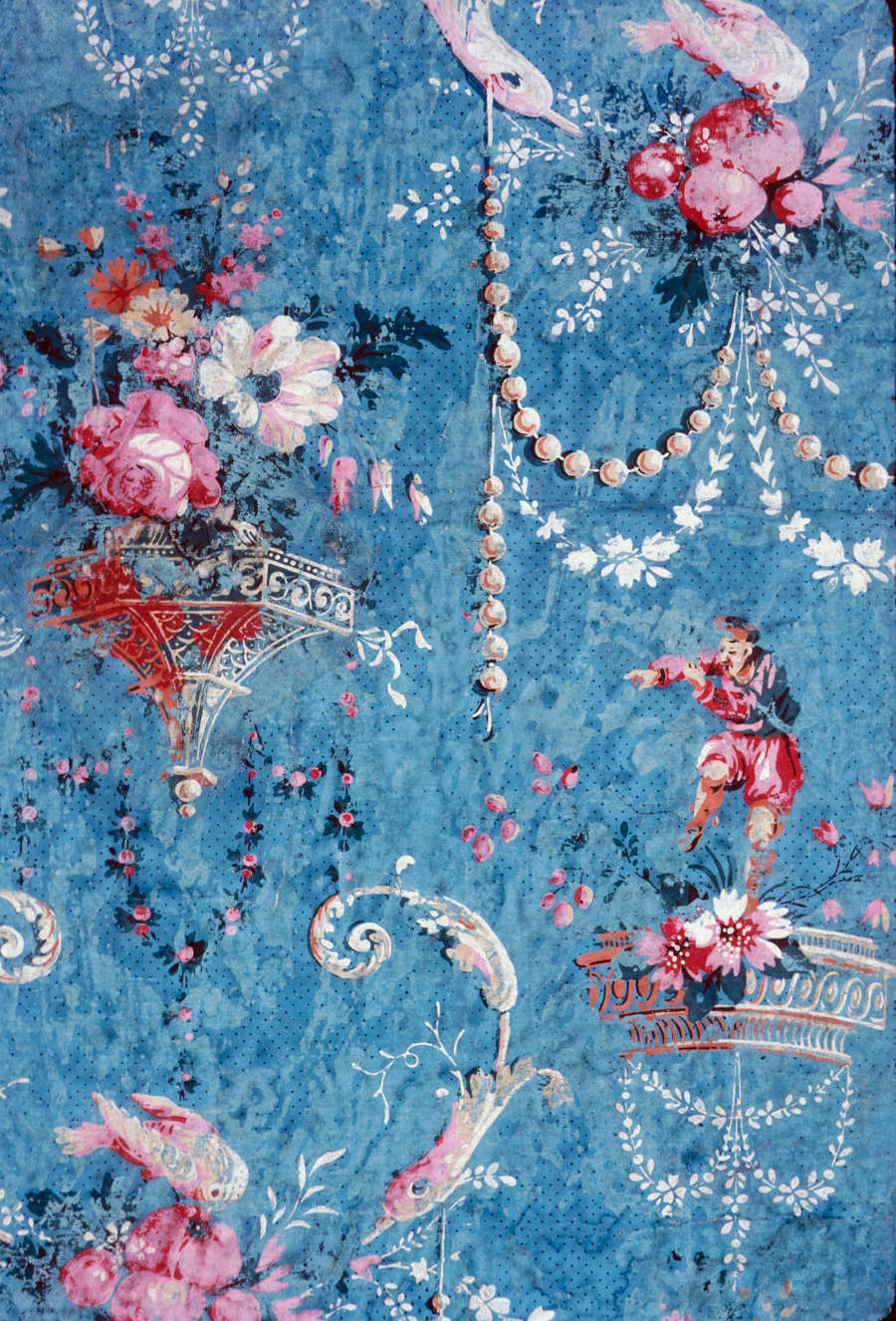 Blue, aged wallpaper with repeating white birds on top of pink flowers, a standing jester, and colorful flower bouquets in elaborate free-standing vases. Winding flower garlands surround the empty space