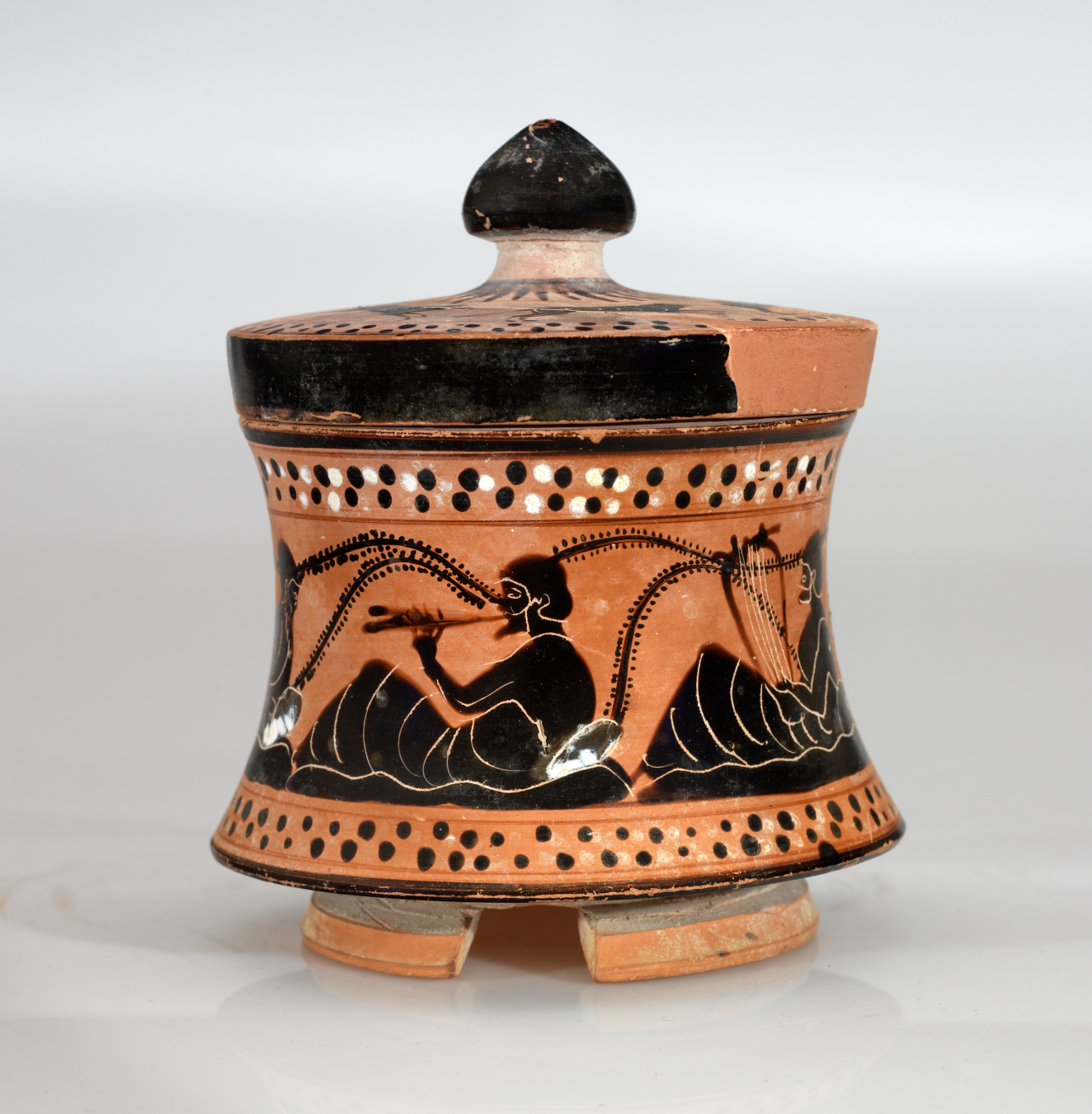 A small, flared, terracotta container with a black-glazed lid, adorned with black figures in flowing robes, holding instruments.