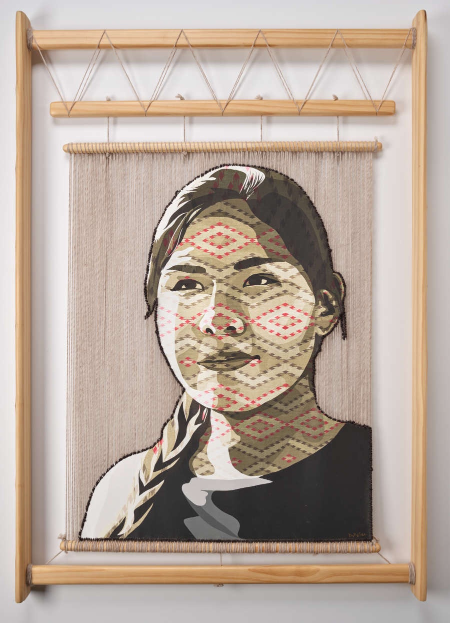 Woven tapestry of a person with braided hair looking off in the distance, displayed on a wooden hand loom. Red and beige geometric patterns sit atop the person's face. 