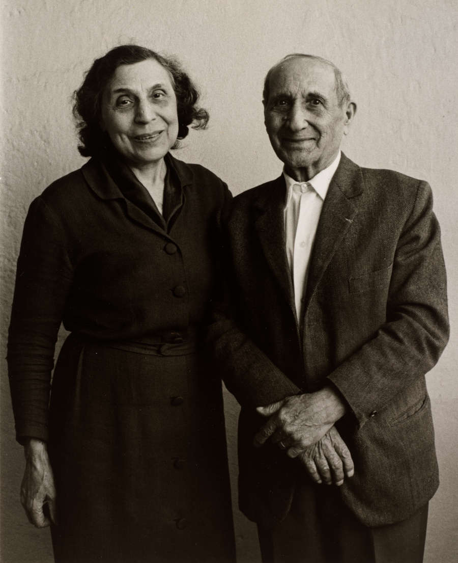 In this black and white photo, an older couple looks sweetly at the camera. Their skin is middle-toned and they are nicely dressed. His large hands are clasped together.