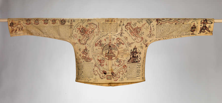 Complete view of the back of a yellowed shirt, densely populated with a symmetrically arranged composition of mythical characters, Buddha, dragons, geometric symbols, grids, and writing in black and red.