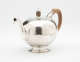 A bulbous silver teapot with a wooden finial and handle connected with decorative silver pieces. There are delicate engraved marks on the body and lid.