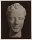 Photograph depicting a sculpture of the head of figure, against a dark background. The head is smooth, despite textured hair. The eyes lack pupils, giving the face a blank look.