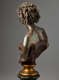 Rear-left angle view of a bronze bust highlighting hair in twists and sculpted, curved shoulder blades exposed above a dark strapless top cut off diagonally and mounted on a gold base.