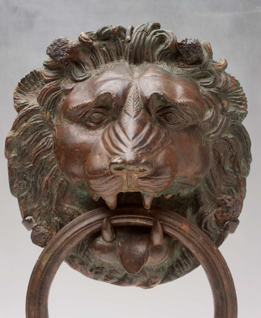 Close-up of the bronze lion’s head biting a circular handle, its tongue outstretched. Its wide open eyes gaze towards the viewers, and ridges form the nose and whiskers.