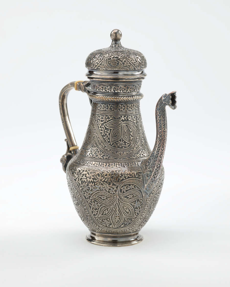 A silver coffee pot with detailed delicate decorations, handle with a lid hinged to it, and spout.