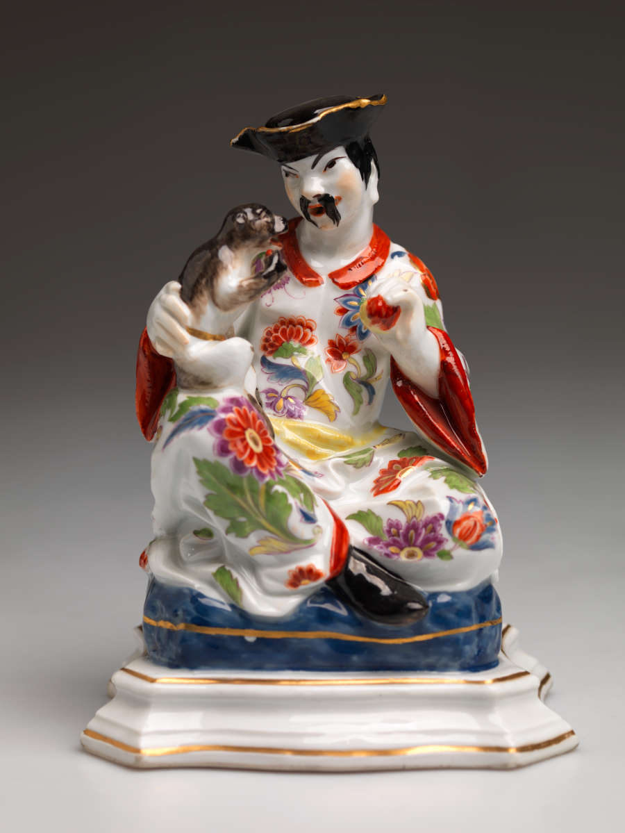 Sculpture of a caricatured Chinese figure wearing a floral garment and black hat. The figure holds a small animal on their lap.