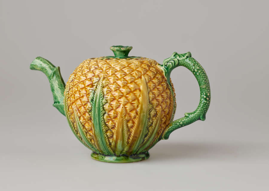 A teapot in the shape of a pineapple. Handle, foot, lid, and spout are green.