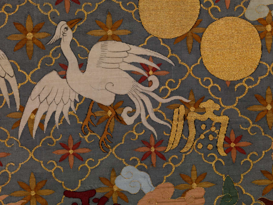 Detail of the robe, featuring a white bird beside two golden circles. The background is dark with a golden diagonal grid, within each square is a floral motif.
