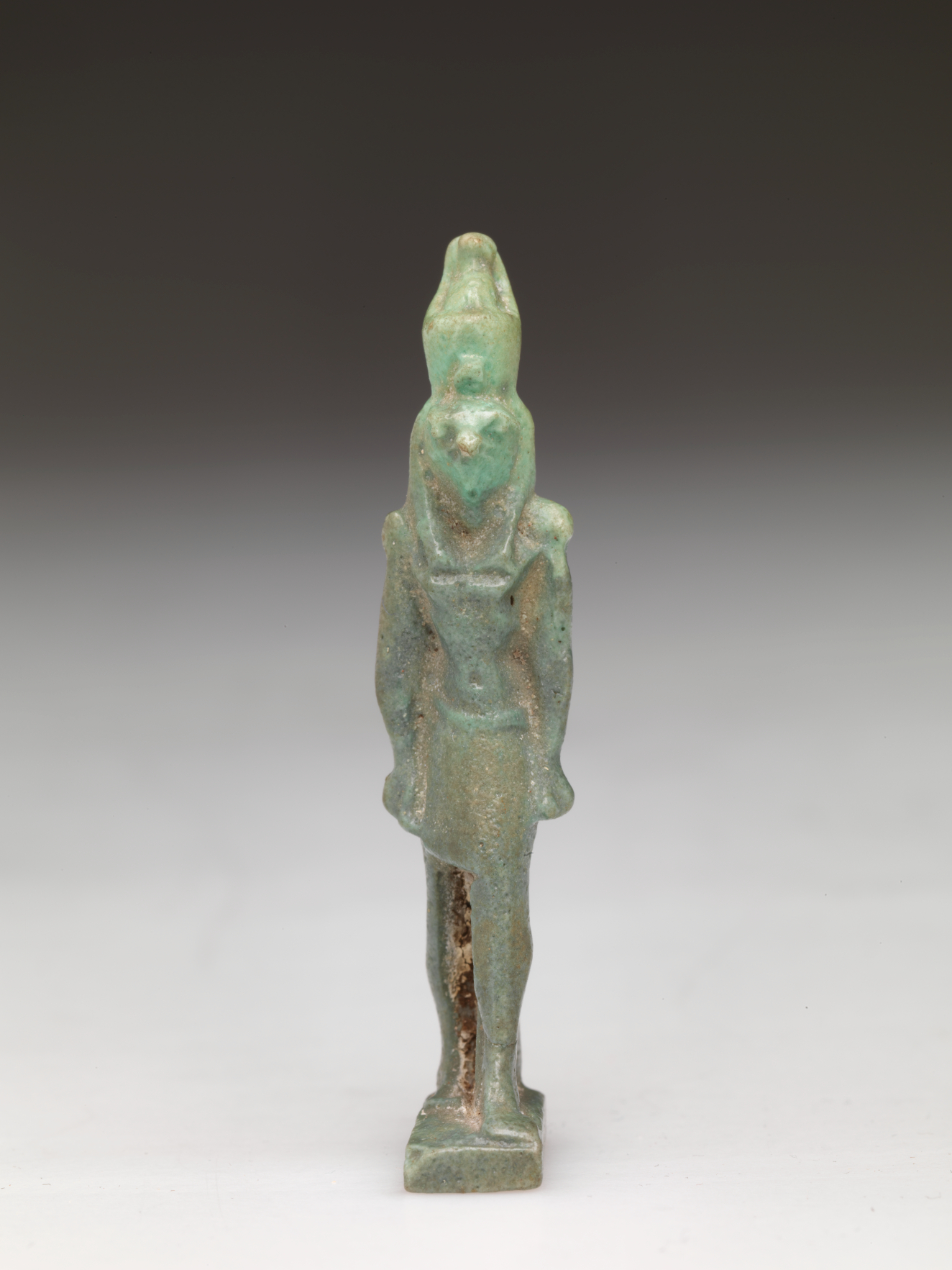 Standing light-jade and brown sculpture of a standing person with a tall head, narrow torso, and straight arms. The left leg has brown specks on it while the headdress is light and translucent.