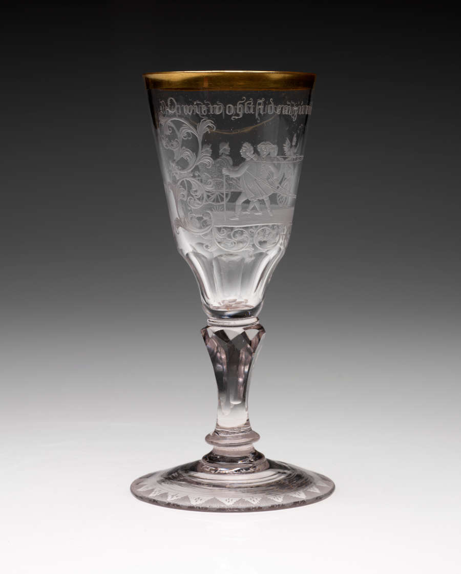 A clear stemmed glass with a rounded foot and thick gold rim. An image of figures walking in a group with words above them are etched in the sides.