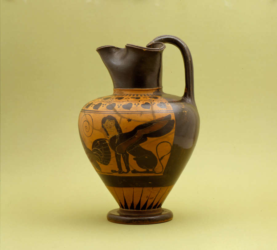 Tall onion-shaped jug with a pinched mouth forming a spout, and a handle. The black and orange jar is decorated with natural and geometric patterns and illustrations of a sphynx.