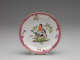 A white plate with a gilded and pink edge and floral patterns. Center design features two birds perched on leafy branches.

