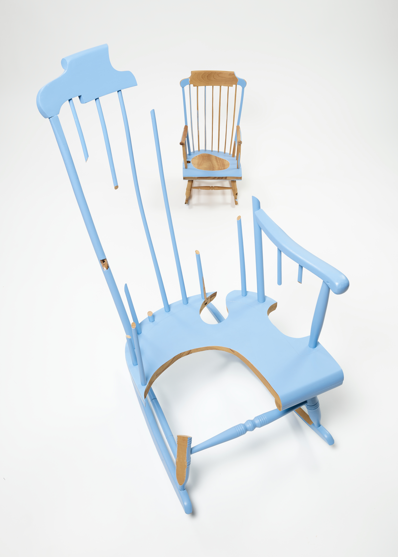 Foreground shows a light blue rocking chair with missing parts, curved cutouts, and exposed wood edges. Behind it sits a smaller natural-wood chair, with partially painted light blue sections.